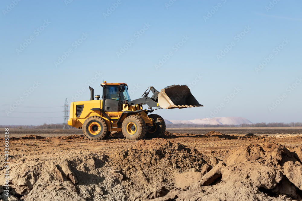 Fototapeta premium Bulldozer or loader moves the earth at the construction site against the blue sky. An earthmoving machine is leveling the site. Construction heavy equipment for earthworks.