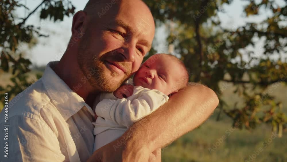 Infant is in father's arms, outdoors while sunset. Happy smiling dad is looking on her newborn baby. Father with newborn son. Happiness: daddy and child. Concept of happy parenthood. Infant is in father's arms, outdoors while sunset. Happy smiling dad is looking on her newborn baby. Father with newborn son. Happiness: daddy and child. Concept of happy parenthood.