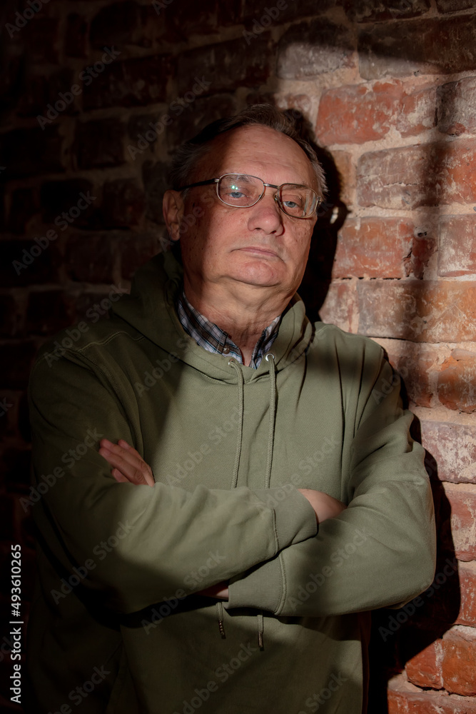 Serious senior male in glasses stand next to brick wall with light from window Serious senior male in glasses stand next to brick wall with light from window
