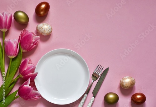 Easter composition.  White plate with cutlery, tulip flowers bouquet and easter eggs on pink background. Top view with copy space. Flat lay. Template for menu