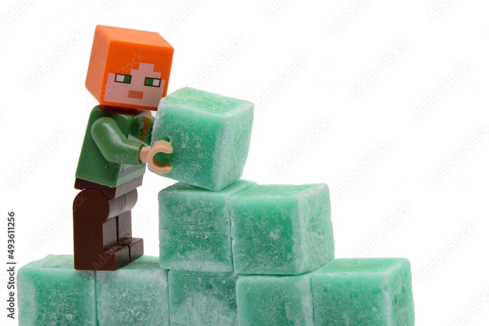 Naklejka premium LEGO Minecraft figure of Alex is building some wall from green coloured scented stearine wax cubes, white background.