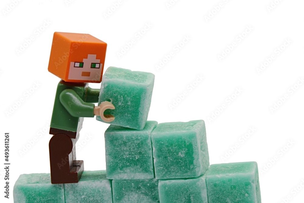 Fototapeta premium LEGO Minecraft figure of Alex is building a wall from green coloured scented stearine wax cubes, possibly of mint or eucalyptus scent, white background