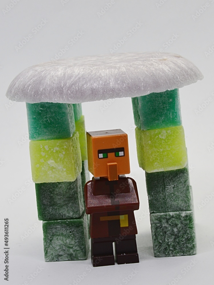 Fototapeta premium LEGO Minecraft figure of villager standing under the roof made of melted scented pink wax, supported by stearine wax cubes of various green colours.