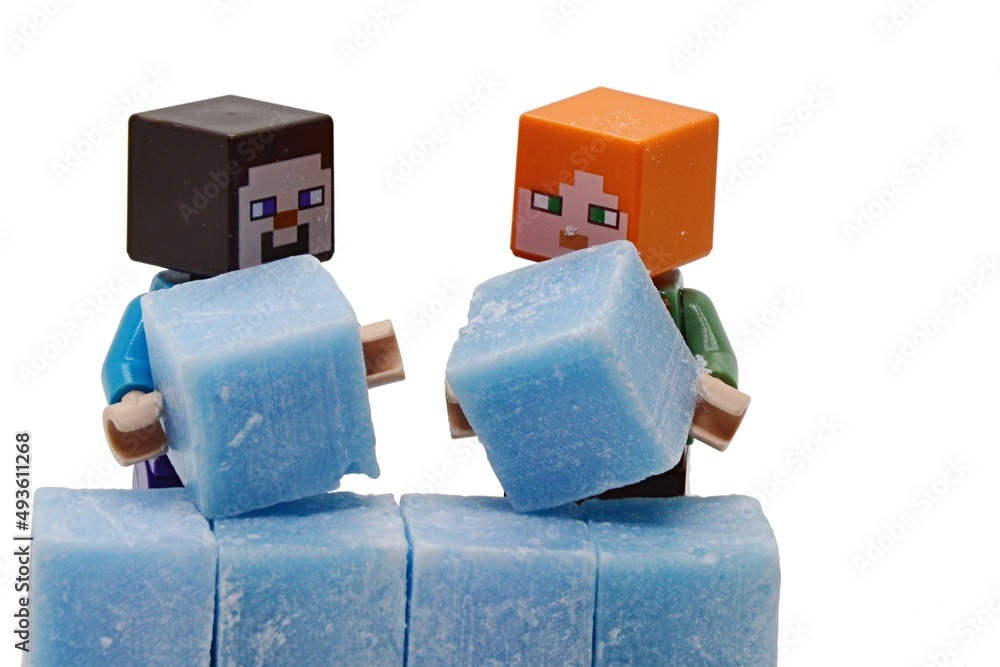 Fototapeta premium LEGO Minecraft figures of Steve and Alex building together a wall of light blue scented stearine wax cubes, white background