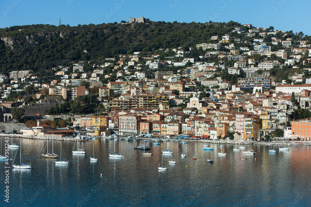 Blue sea with small luxury yachts and boats with cute European French village on a hill at background on sunny summer day. Aerial view of Mediterranean sea in french riviera. High quality photo