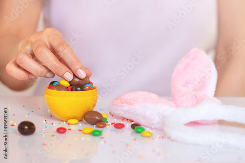 Faceless woman at table. Female hand takes chocolate candy from yellow Easter egg. Funny bunny ears and scattered sweets on surface.