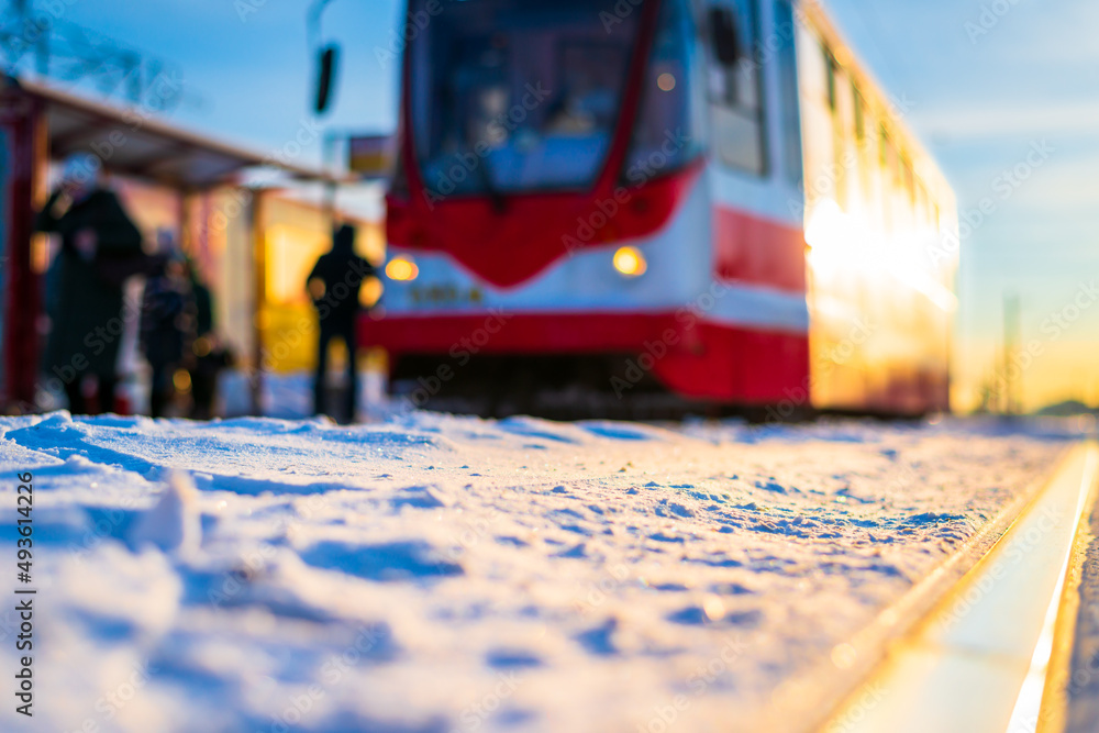 Tram stop with people. Sunrise on a winter morning. The tram is moving forward. Snow-covered road. Focus on rail. Close up view from ground level.