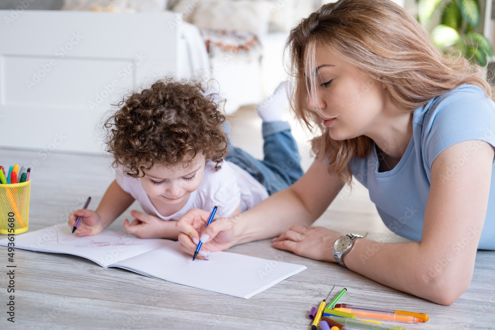 young mother and curly child girl drawing with colored markers lying on floor room at home young mother and curly child girl drawing with colored markers lying on floor room at home