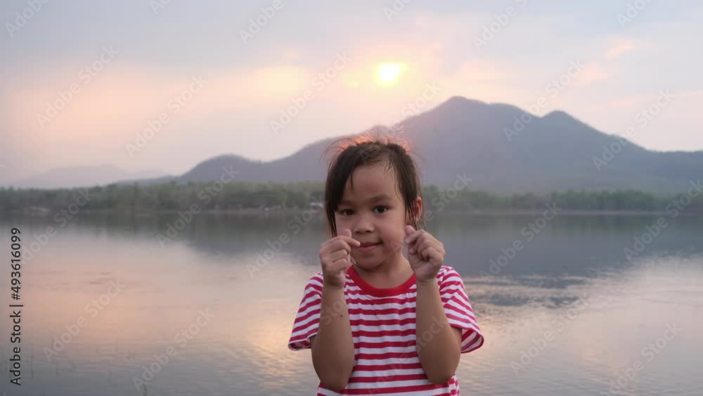 Smiling cute little girl doing mini heart gesture with her hands and looking at the camera during the sunset by the lake. Travel on summer vacation with family. Smiling cute little girl doing mini heart gesture with her hands and looking at the camera during the sunset by the lake. Travel on summer vacation with family.