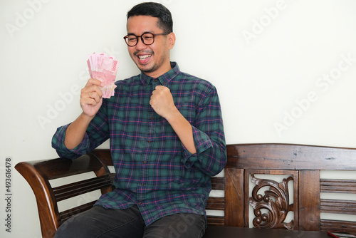 Adult Asian man sitting in a bench showing happy expression while looking to paper money that he hold photo