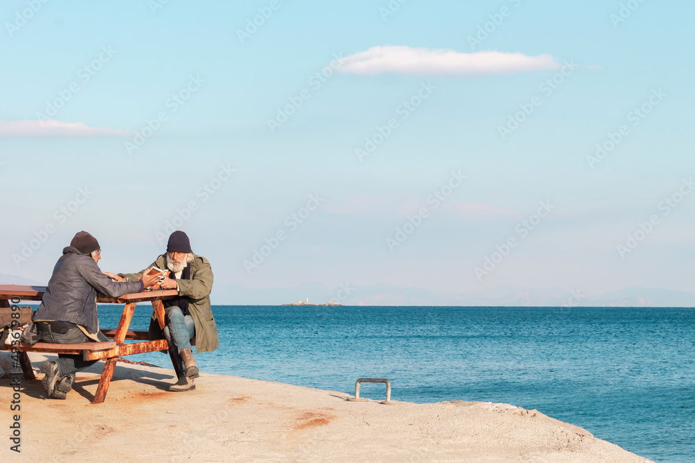 Old men sits on a bench by the sea. One man suggesting a book the other man.	
