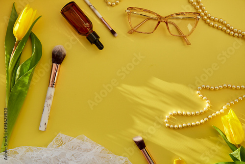 Pastel feminine flat lay with pearls, makeup brushes, glasses, serum dropper and lace. Yellow tulips with trendy shadows. Beauty card concept. Copy space photo