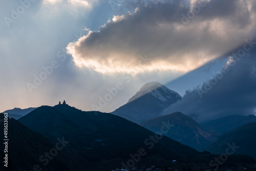 Fototapeta Naklejka Na Ścianę i Meble -  Distant view on Gergeti Trinity Church in Stepansminda, Georgia. The church is located the Greater Caucasian Mountain Range.Clouds are covering the snow-capped Mount Kazbegi in the back.Sunrise,sunset