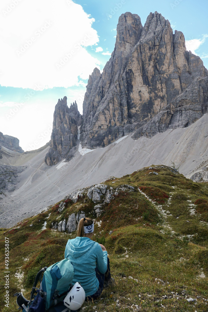 A woman in blue pullover resting under the foot of high Italian Dolomites. There are many sharp peaks in front of her. She is recharging before the further climb. Lots of lose stones and landslides.