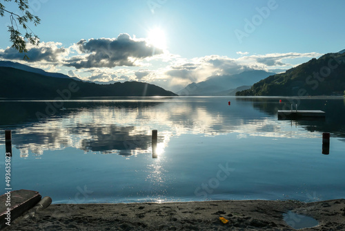 A sunset by Millstaetter lake in Austria. The lake is surrounded by high Alps. Calm surface of the lake reflecting the sunbeams. The sun sets behind the mountains. A bit of overcast. Natural beauty