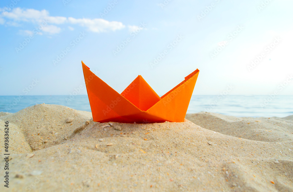 Bright orange paper boat in yellow sand on background of sea waves and blue sky on sunny summer day. Colored ship close-up. Concept adventures travelling vacation holiday rest tourism voyager dream Bright orange paper boat in yellow sand on background of sea waves and blue sky on sunny summer day. Colored ship close-up. Concept adventures travelling vacation holiday rest tourism voyager dream