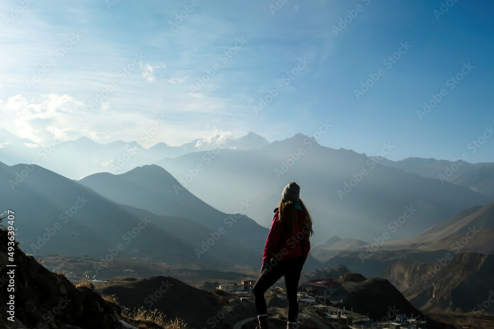 Fototapeta premium A girl wearing pink jacket standing on a top of a Himalayan mountain and enjoying the misty range spreading in front of her. Muktinath, Nepal. The sunlight is nicely marked. Freedom and achievement.