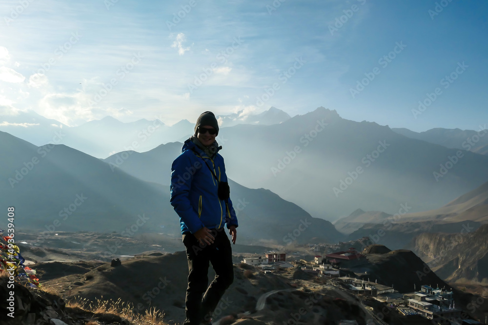 A man in hiking outfit standing on top of a mountain with the endless view on Himalayan chains. There is a small village in the valley. Golden hours in Muktinath, Nepal. Achievement