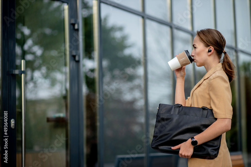 Confident businesswoman walking around the city talking on the phone with a wireless headset and smart watch. Young woman talking on the phone and drinking coffee while walking.