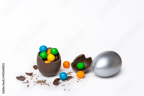 Cracked chocolate easter egg with colorful small round sweets and chocolates and a whole painted silver egg on a white background, copy space. Easter concept.