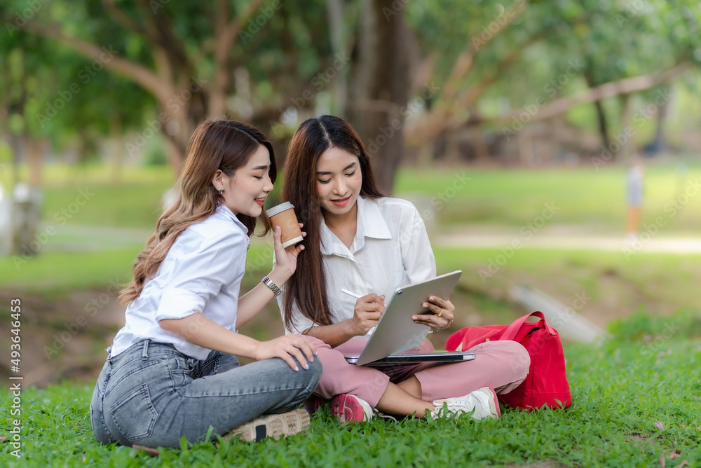 Two Asian female students talking and using a tablet sitting in the park on university. Two Asian female students talking and using a tablet sitting in the park on university.