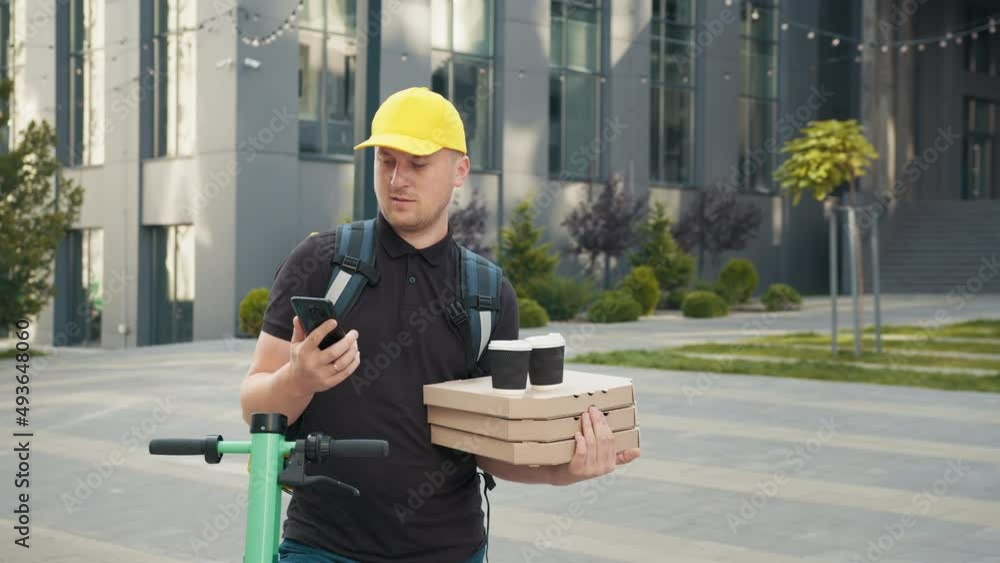 Portret Young courier, pizza and coffee delivery man with thermal backpack and phone on a E-Scooter. Fast transport express home delivery. Online order. Delivery man in cap looking at smartphone. Portret Young courier, pizza and coffee delivery man with thermal backpack and phone on a E-Scooter. Fast transport express home delivery. Online order. Delivery man in cap looking at smartphone.