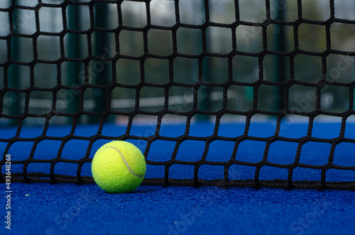 Paddle tennis ball next to the net of a blue artificial grass paddle tennis court.