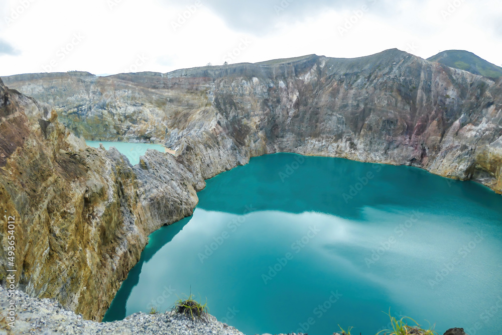 Fototapeta premium View on the Kelimutu volcanic crater lakes in Flores, Indonesia. Lakes are shining with many shades of turquoise and blue. Sun shines through clouds. Barren and sharp slopes of the volcanic crater
