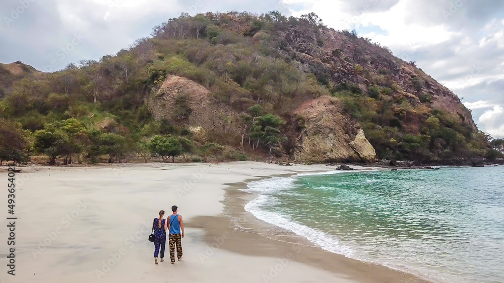A couple walking on an idyllic Koka Beach. Hidden gem of Flores, Indonesia. Couple is enjoying their romantic escape. Waves gently washing the shore. There are hills in the back. Happiness and love A couple walking on an idyllic Koka Beach. Hidden gem of Flores, Indonesia. Couple is enjoying their romantic escape. Waves gently washing the shore. There are hills in the back. Happiness and love