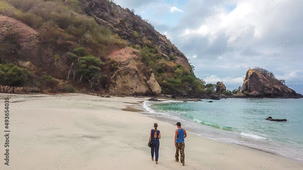 A couple walking on an idyllic Koka Beach. Hidden gem of Flores, Indonesia. Couple is enjoying their romantic escape. Waves gently washing the shore. There are hills in the back. Happiness and love A couple walking on an idyllic Koka Beach. Hidden gem of Flores, Indonesia. Couple is enjoying their romantic escape. Waves gently washing the shore. There are hills in the back. Happiness and love