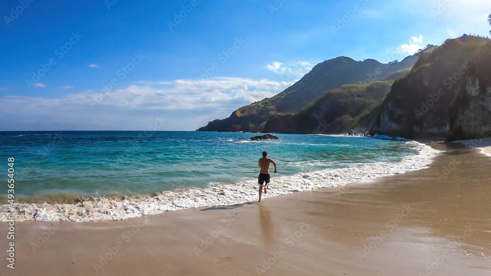 A man running along the shore, chasing the waves on idyllic Koka Beach. Hidden gem of Flores, Indonesia. He is having a lot of fun, collecting happy moments. Adventure and discovering while travelling A man running along the shore, chasing the waves on idyllic Koka Beach. Hidden gem of Flores, Indonesia. He is having a lot of fun, collecting happy moments. Adventure and discovering while travelling