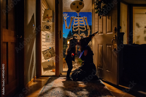 A mom dressed as a witch and a daughter dressed as a cat kiss in front of a front door that is decorated with a skeleton and other halloween decor