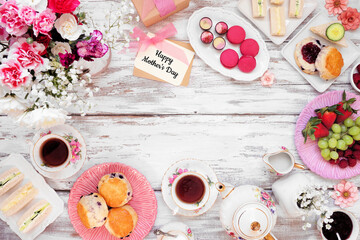 Mother's Day tea frame over a white wood background. Greeting card, flowers, vintage tea set, finger sandwiches, chocolates, desserts and fruit. Copy space.