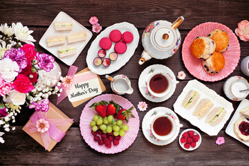 Mother's Day tea table scene against a dark wood background. Greeting card, flowers, gift, vintage tea set, finger sandwiches, chocolates, desserts and fruit.