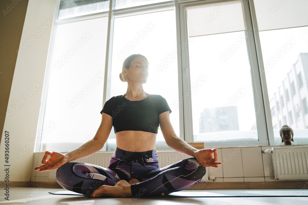 A woman meditates with her eyes closed. Sit in a yoga pose, silence in thoughts, calm the breath, hatha yoga, a person practices meditation, light in the window, a sunny day.