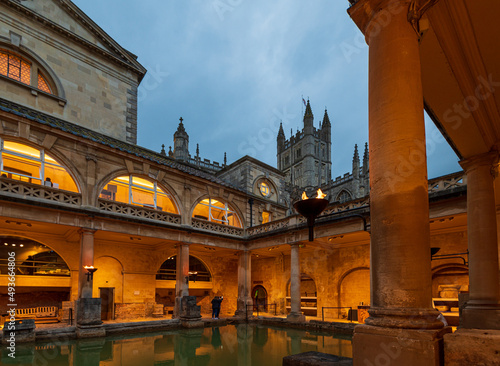 Bath town, UK. Evening sightseeing of restored in Victorian times ancient Roman Baths in the fire light of wall sconces. In the background visible tower of Abbey Church of Saint Peter and Saint Paul.