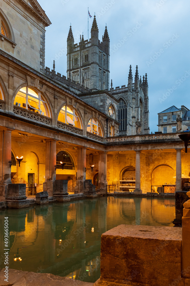 Bath town, UK. Evening sightseeing of restored in Victorian times ancient Roman Baths in the fire light of wall sconces. In the background visible tower of Abbey Church of Saint Peter and Saint Paul.