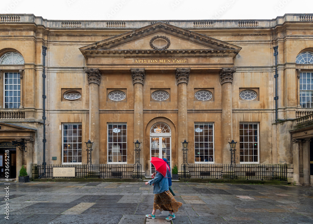 City of Bath, UK. People with red umbrella walking in front of restored in Victorian times ancient Roman Baths.  The Pump Room. Translation from old Greek: Water is Best.