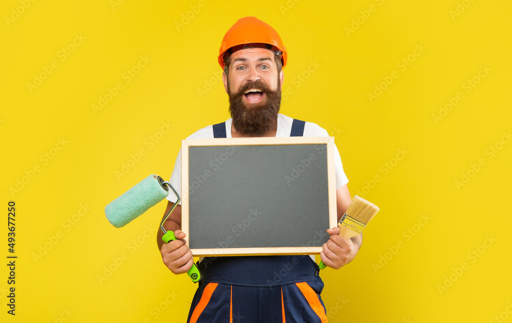 happy man painter in helmet and work clothes hold paint roller brush and blackboard with copy space happy man painter in helmet and work clothes hold paint roller brush and blackboard with copy space