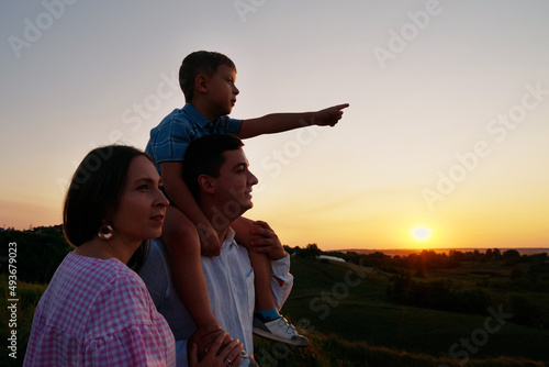 Family standing on hill in countryside at sunset. Arc shot little boy sitting on shoulders of father and pointing at something in the distance, mother standing beside. Concept of nature