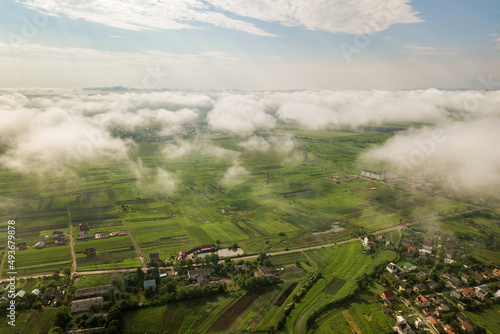 Aerial view of white clouds above a town or village with rows of buildings and curvy streets between green fields in summer. Countryside landscape from above. © bilanol