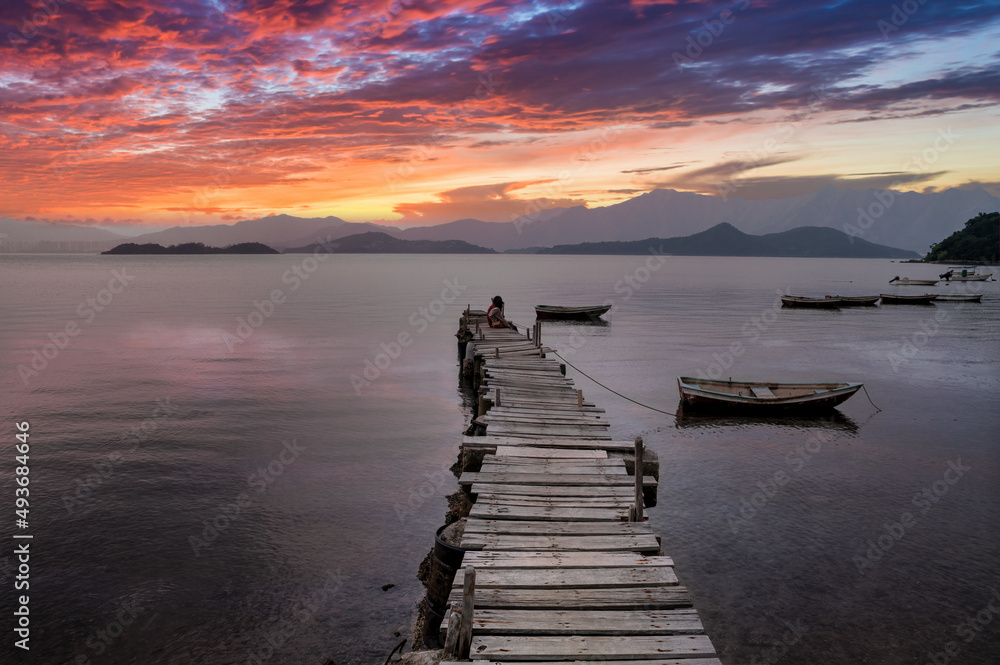 Sunset over Tolo Harbour with two youths sitting on a rickety wooden pier, Ma On Shan, Hong Kong. 