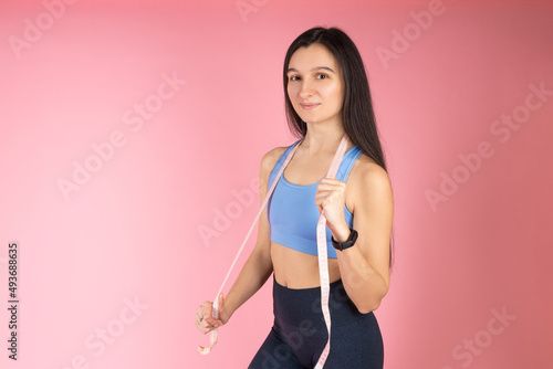 The trainer holds in his hands a measuring tape to measure the body. Attractive brunette woman in fashionable sportswear on pink background. Healthy lifestyle concept © mdyn