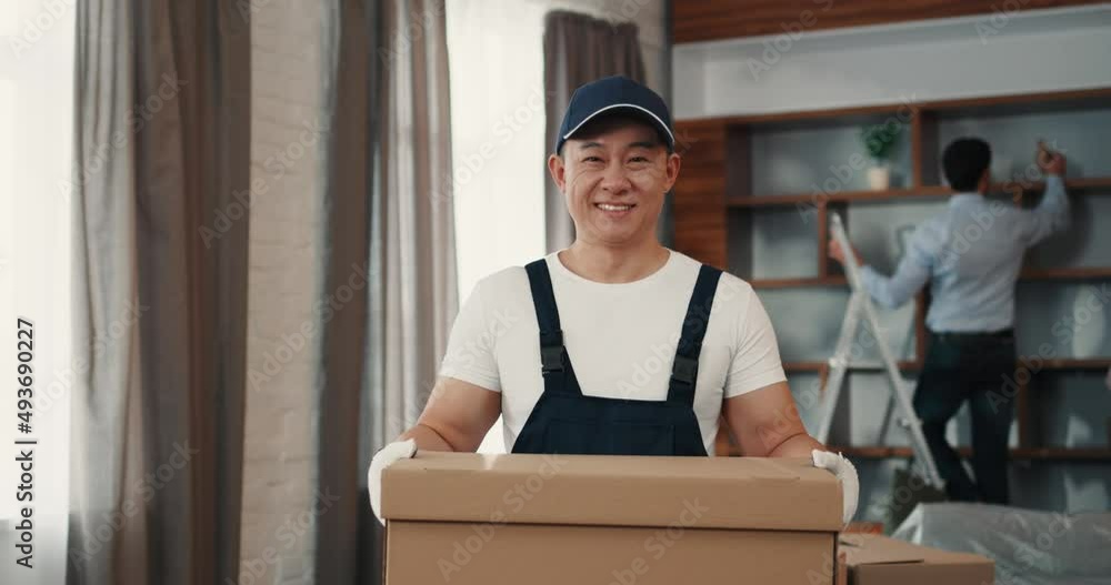Portrait of male worker of relocation service, dressed in uniform, holding cardbox and smiling at came in apartment Portrait of male worker of relocation service, dressed in uniform, holding cardbox and smiling at came in apartment