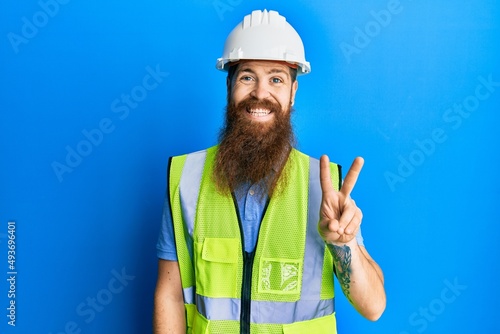 Redhead man with long beard wearing safety helmet and reflective jacket showing and pointing up with fingers number two while smiling confident and happy.