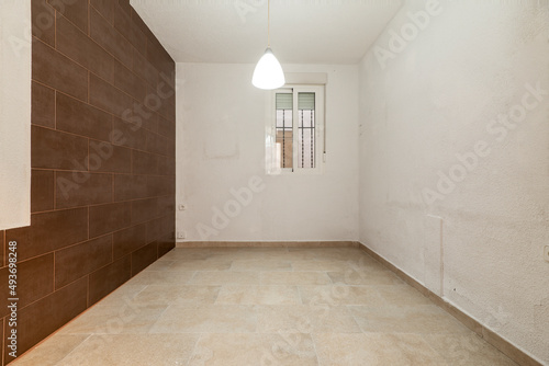 Empty room with a wall covered in dark brown tiles  a pine wood window and door  glass pavers on the walls  an aluminum radiator and a stoneware floor