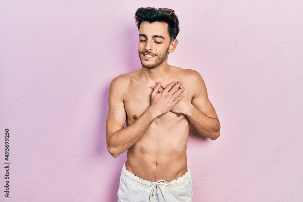 Fototapeta premium Young hispanic man wearing swimwear shirtless smiling with hands on chest with closed eyes and grateful gesture on face. health concept.