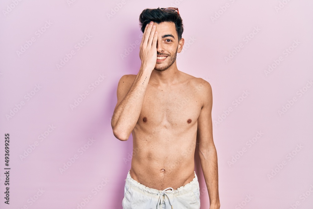 Fototapeta premium Young hispanic man wearing swimwear shirtless covering one eye with hand, confident smile on face and surprise emotion.
