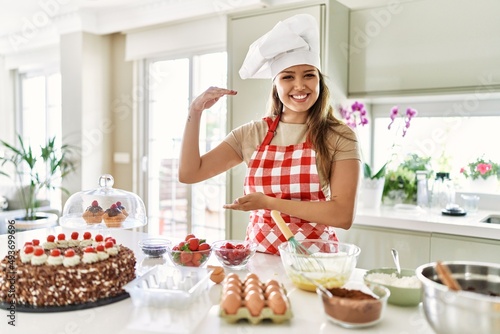 Beautiful young brunette pastry chef woman cooking pastries at the kitchen gesturing with hands showing big and large size sign, measure symbol. smiling looking at the camera. measuring concept.