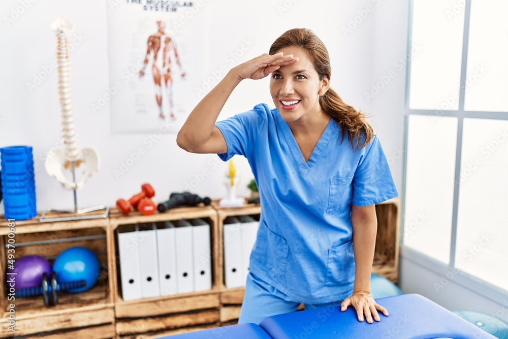 Middle age hispanic physiotherapist woman working at pain recovery clinic very happy and smiling looking far away with hand over head. searching concept. Middle age hispanic physiotherapist woman working at pain recovery clinic very happy and smiling looking far away with hand over head. searching concept.